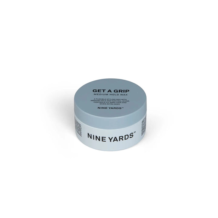 Apvali, plokščia, skalūno mėlyna NINE YARDS Get A Grip Medium Hold Wax 100ml dėžutė, kurios šone juodomis raidėmis užrašytas produkto pavadinimas ir aprašymas. Produktas baltame fone.